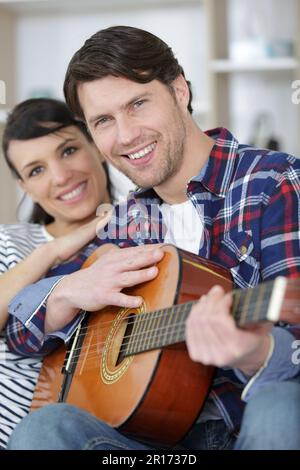 Ein Paar, das auf dem Sofa kuschelt, während ein Mann Gitarre spielt Stockfoto