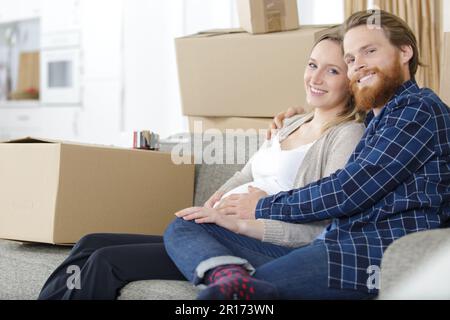 Ein Paar, das ein Baby erwartet, saß auf dem Sofa, umgeben von Pappkartons Stockfoto
