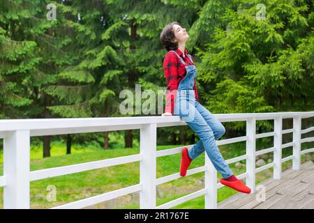 Glückliche junge Frau in blauem Jeansanzug und rotem Hemd sitzt auf einem Holzzaun auf der Terrasse im Hinterhof, vor dem Hintergrund eines grünen Rasen und Waldes an einem sonnigen Tag Stockfoto