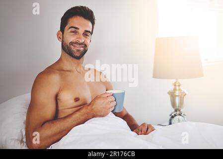 Es ist der einzige Weg, aufzuwachen. Abgeschnittenes Porträt eines gutaussehenden jungen Mannes, der eine Tasse Kaffee genießt, während er zu Hause in seinem Bett sitzt. Stockfoto