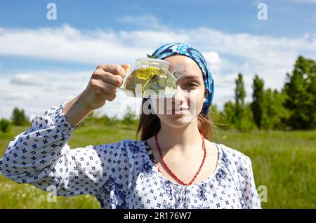 Frau mit transparenter Teekanne und Kräutertee. Sammelt frische grüne Kräuter, ernten und machen gesunden Tee. Verschwommenes Bild, selektiver Fokus Stockfoto