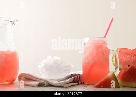 Frisch zubereitetes Wassermelonengetränk mit zerstoßenem Eis auf dem Tisch mit geschnittenen Früchten, zerstoßenem Eis und Krug. Vorderansicht. Horizontale Zusammensetzung. Stockfoto