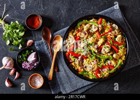 Hähnchenpilzpaella mit süßer Paprika, grünen Erbsen und Gewürzen in schwarzer Schüssel auf Betontisch mit Holzlöffeln und Gewürzen, horizontale Ansicht von Stockfoto