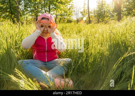 Ein fröhliches Mädchen sitzt im dicken grünen Gras und legt ihre Handflächen in Form einer Brille an ihr Gesicht. Machen Sie einen Spaziergang im Stadtpark Stockfoto