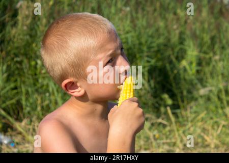 Wunderschöner weißer Junge, der gekochten Mais in der Natur isst Stockfoto