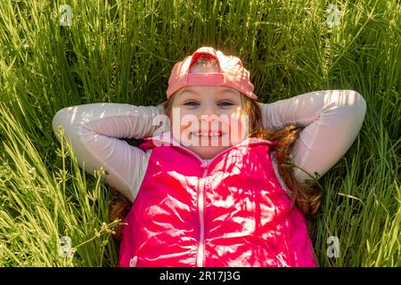 Ein fröhliches Mädchen liegt auf dem Rücken im dicken grünen Gras. Machen Sie einen Spaziergang im Stadtpark Stockfoto