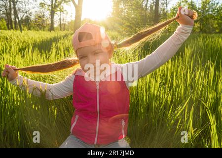 Ein fröhliches Mädchen sitzt im dicken grünen Gras und hält ihre Zöpfe mit den Händen, vor dem Hintergrund des Sonnenuntergangs. Machen Sie einen Spaziergang im Stadtpark Stockfoto