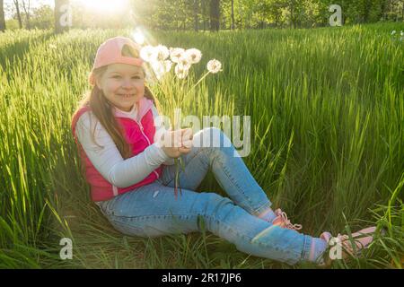 Ein fröhliches Mädchen mit einem Blumenstrauß von Löwenzahn sitzt im dicken grünen Gras vor dem Hintergrund des Sonnenuntergangs. Machen Sie einen Spaziergang im Stadtpark Stockfoto