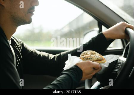 Ein Mann mit Frühstücks-Bagel, der auf dem Weg zur Arbeit im Auto fährt Stockfoto