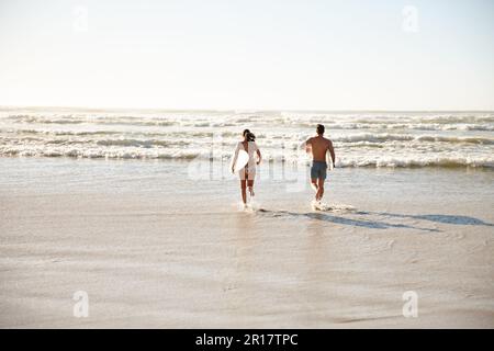 Perfekter Sommer. Rückansicht eines jungen Surferpaares, das zusammen ins Meer rennt. Stockfoto