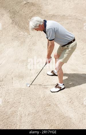 Steckt in der Sandfalle fest. Ein älterer Mann, der versucht, seinen Ball aus der Sandfalle auf dem Golfplatz zu schlagen. Stockfoto