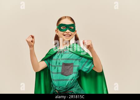 Mächtiges Mädchen in grünem Superheldenkostüm mit Umhang und Maske im Gesicht, lächelt und steht mit geballten Fäusten, während sie den World Child Protectio feiert Stockfoto
