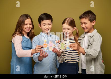 Lächelnde Kinder interrassischer Kinder in lässiger Kleidung mit Freunden, die gezeichnete Papierfiguren halten, während sie den Tag des Kinderschutzes auf Khaki backgro feiern Stockfoto