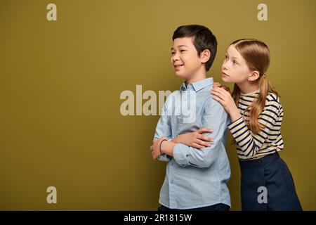 Rothaariges Mädchen in gestreifter Bluse umarmt lächelnde asiatische Freunde, die während der Feier zum Kinderschutztag auf Khaki-Hintergrund wegschauen Stockfoto