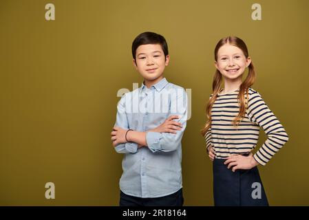 Lächelndes rothaariges Mädchen, das Hände an der Hüfte hält und in die Kamera schaut, während ein asiatischer Freund während der Feier zum Kinderschutztag auf Khaki Back die Arme kreuzte Stockfoto