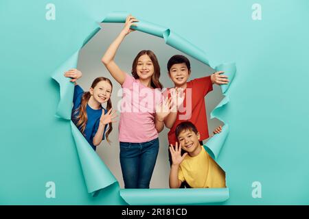 Lächelnde Kinder unterschiedlicher Rassen in lässiger Kleidung, die während des internationalen Kindertags mit den Händen vor der Kamera schwenken, hinter einem Loch in blauem Papierhintergrund Stockfoto