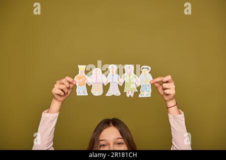 Zugeschnittener Blick auf ein junges Mädchen, das handgezeichnete Papierfiguren über dem Kopf hält und während der Kinderschutzfeier auf Khaki Bac in die Kamera schaut Stockfoto