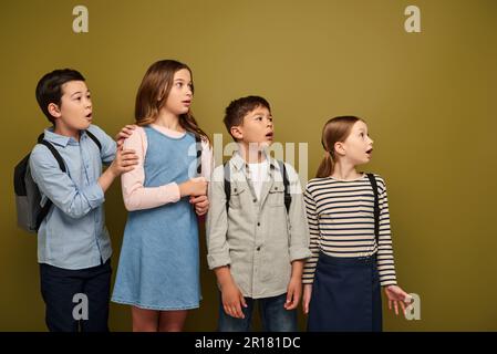 Schockierte multiethnische Kinder in lässiger Kleidung mit Rucksäcken, die wegschauen und während der Kinderschutzfeier auf khak den Mund öffnen Stockfoto