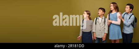 Überraschte multikulturelle Schulkinder mit Rucksäcken, die während der internationalen Kinderschutzfeier auf Khaki-Hintergrund mit Copy Spa abblickten Stockfoto