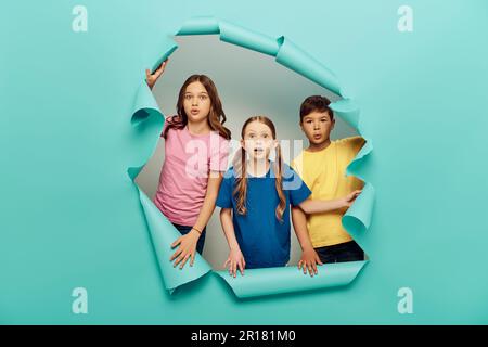 Schockierte multiethnische Kinder in farbenfrohen T-Shirts, die in die Kamera schauten, während sie den Tag des Kinderschutzes hinter dem Loch in blauem Papierhintergrund feierten Stockfoto