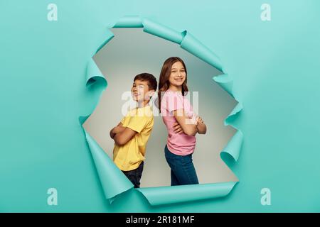 Lächelnde Kinder interrassischer Herkunft in bunten T-Shirts, die die Arme kreuzen, während sie sich Rücken an Rücken zurücklehnen und den Tag des Kinderschutzes hinter dem Loch in BL feiern Stockfoto