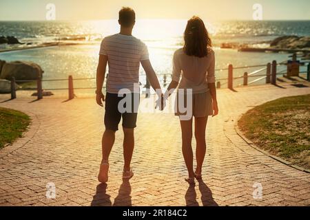 In den Sonnenuntergängen zusammenhalten... das ist wahre Liebe. Rückblick auf ein junges Paar, das im Sommer den Sonnenuntergang an der Küste beobachtet. Stockfoto