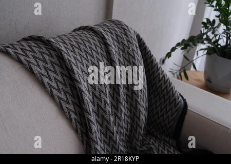 Weißes Sofa mit einer Decke, Decke mit Zickzack-Muster auf einem weißen Sofa mit gelber Pflanze. Stockfoto