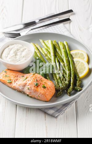 Gesundes Essen gebackenes Lachsfilet mit Spargel serviert mit Tartar Sauce und Zitrone Nahaufnahme auf einem Teller auf dem Tisch. Vertikal Stockfoto