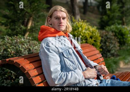 Ernsthaft fokussierter deutscher junger Mann, der auf einer Holzbank im Park sitzt und an sonnigen Tagen in die Kamera schaut Stockfoto