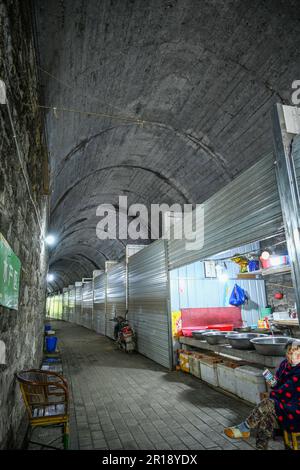 CHONGQING, CHINA - 8. MAI 2023 - das am 8. Mai 2023 aufgenommene Video zeigt einen Lebensmittelmarkt in einem verlassenen Eisenbahntunnel in Chongqing, China. In diesem hundert Meter langen Tunnel sind mehr als 20 Stände nacheinander angeordnet. Die Verkaufsstände sind in feste Verkaufsstände und provisorische Verkaufsstände unterteilt, und die festen Verkaufsstände sind in Fleischbereiche und Bereiche für Wasserprodukte unterteilt. Es wird davon ausgegangen, dass der Tunnel Teil der ehemaligen Eisenbahnlinie des Spezialstahlwerks Chongqing ist, die eine Tunnellänge von etwa 150 Metern hat. Nach dem Bankrott des Spezialstahlwerks Chongqing wurden die einst befahrenen Tunnel allmählich still, A Stockfoto