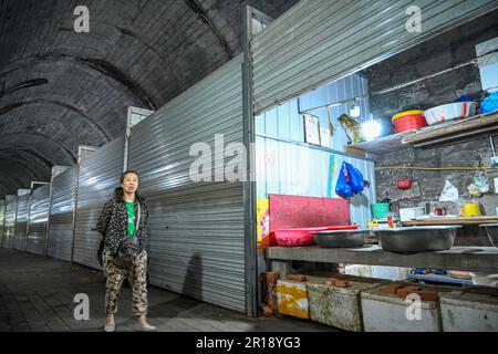 CHONGQING, CHINA - 8. MAI 2023 - das am 8. Mai 2023 aufgenommene Video zeigt einen Lebensmittelmarkt in einem verlassenen Eisenbahntunnel in Chongqing, China. In diesem hundert Meter langen Tunnel sind mehr als 20 Stände nacheinander angeordnet. Die Verkaufsstände sind in feste Verkaufsstände und provisorische Verkaufsstände unterteilt, und die festen Verkaufsstände sind in Fleischbereiche und Bereiche für Wasserprodukte unterteilt. Es wird davon ausgegangen, dass der Tunnel Teil der ehemaligen Eisenbahnlinie des Spezialstahlwerks Chongqing ist, die eine Tunnellänge von etwa 150 Metern hat. Nach dem Bankrott des Spezialstahlwerks Chongqing wurden die einst befahrenen Tunnel allmählich still, A Stockfoto