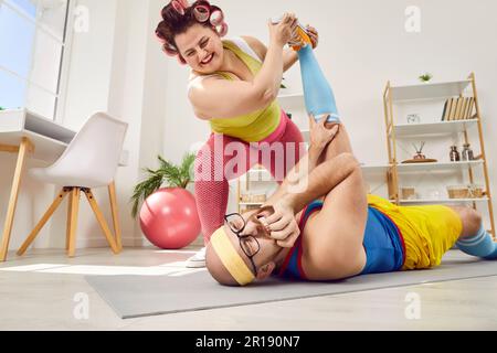 Lustige, übergewichtige junge Frau lässt ihren Sporttrainer schmerzhafte Beindehnung üben Stockfoto