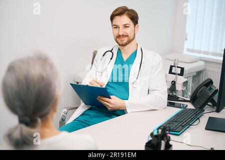 Ein angenehmer junger Arzt kommuniziert mit einer älteren Frau im Therapieraum Stockfoto