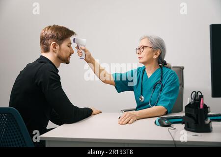 Bärtige Patientin, eine Ärztin misst die Temperatur mit einem modernen Thermometer Stockfoto