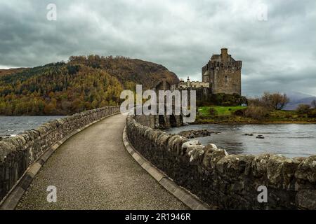 Alte schottische Burg von Eilean Donan in der Nähe von Dornie führt eine Steinbrücke zum Schloss und ein Fluss verläuft darunter. Ein Hügel mit Bäumen auf dem Hang Stockfoto