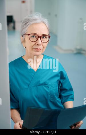Grauhaarige Sanitäterin steht im Krankenhausgang mit MRT-Scan Stockfoto