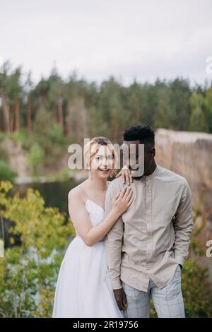 Glückliches gemischtrassiges Paar frisch Vermählte stehen und lachen fröhlich vor dem Hintergrund des Sees und des Waldes. Konzept von Liebesbeziehungen und Einheit zwischen Stockfoto