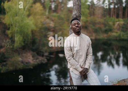 Der junge glückliche afrikaner steht, genießt und träumt mit geschlossenen Augen in der Nähe des Baumes vor dem Hintergrund des Sees und des Waldes während des Ausflugs. Stockfoto