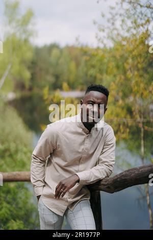 Ein junger glücklicher afrikaner steht auf einer Holzbrücke vor dem Hintergrund von See und Wald während des Ausflugs. Stockfoto