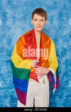 Porträt eines jungen Schwulen, eingewickelt mit einer lgbt-Flagge, der in die Kamera schaut und während der Feierlichkeiten des Pride Month auf einem fleckigen blauen Hintergrund steht Stockfoto