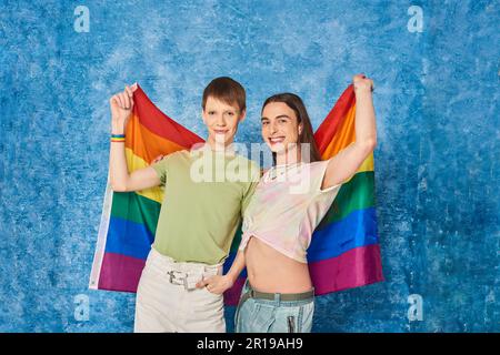 Fröhliche und junge homosexuelle Freunde halten die lgbt-Flagge zusammen und schauen während der Community-Feier im Pride Month auf dem fleckigen blauen Backgr in die Kamera Stockfoto