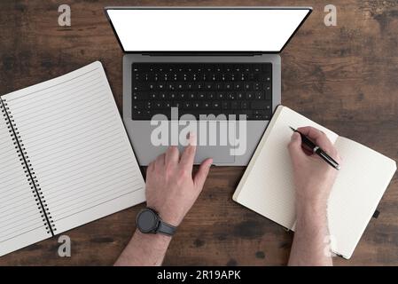 Von oben nach unten: Person, die einen modernen Laptop am Holztisch benutzt und sich auf Notizblock Notizen macht Stockfoto