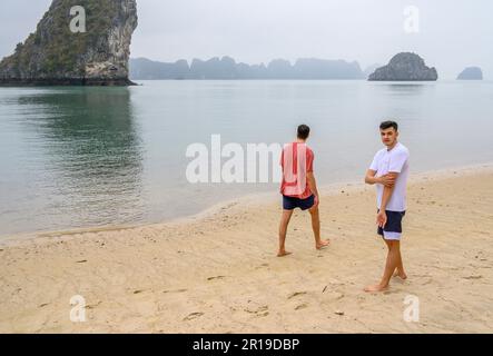 Zwei junge Männer, die an einem Strand vor dem Hintergrund der typischen Kalksteininseln in der Bai TU Long Bay, Halong Bay, Vietnam spazieren. Stockfoto