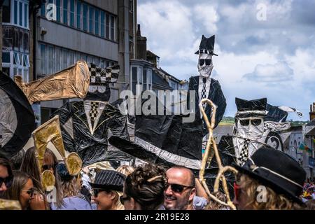 Leute, die große mit- und Papierfiguren von unhöflichen zwei-Ton-Figuren bei der Mazey-Day-Parade beim Golowan Festival in Penzance in Cornwall i dabei hatten Stockfoto