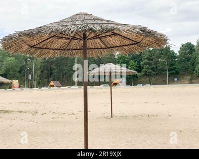 Ein leerer verlassener Strand bei schlechtem Wetter, kalter Herbst in der Nebensaison mit strohgedeckten Sonnenschirmen am blauen Himmel. Stockfoto