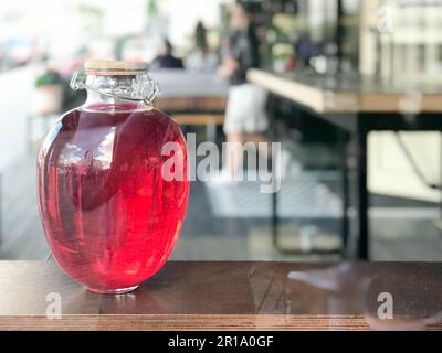 Eine große, wunderschöne drei-Liter-Glasbank mit Schinken, Saft, Trank, einem Getränk mit einer glühenden Flüssigkeit und einem Holzdeckel auf einem verschwommenen Hintergrund auf einem Regal i. Stockfoto