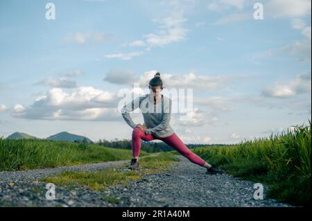 Blick auf eine junge Frau in pinkfarbenen Leggings, die sich dehnt und sich aufwärmt, bevor sie einen Lauf macht. Auf einer Landstraße zwischen grünen Quellfeldern. Stockfoto