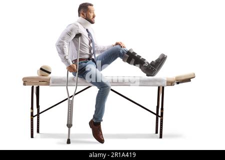 Profilaufnahme eines Verletzten mit orthopädischem Stiefel und Armschlinge auf einem Therapietisch isoliert auf weißem Hintergrund Stockfoto