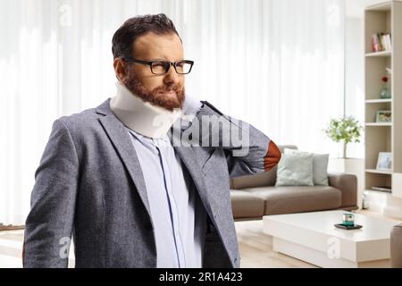 Bärtiger Mann mit Halsband, der seinen schmerzhaften Hals in einem Wohnzimmer zu Hause hält Stockfoto