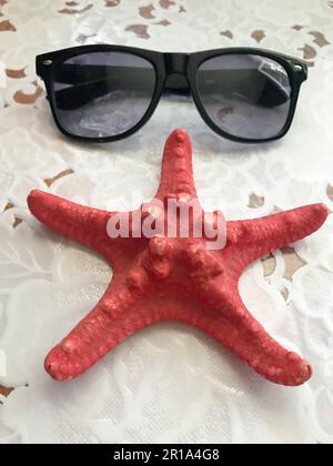 A beautiful red starfish and black sunglasses lie decorated with seashells on a white cloth background. Marine tropical summer background. Stockfoto